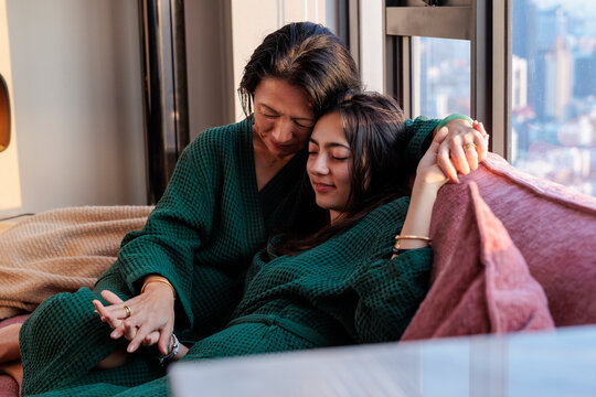 Mother embraces daughter on sofa in robes with peaceful city view