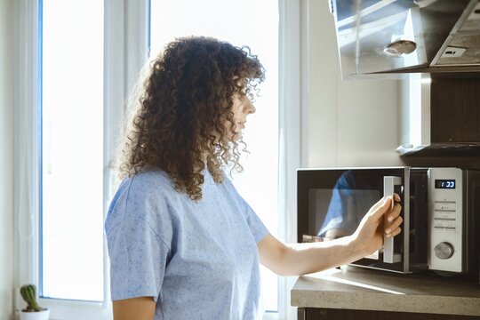 A woman stands by a microwave in a kitchen. She is preparing to heat food. Sunlight enters through the window. The kitchen is tidy with simple decor. There is a plant in the background