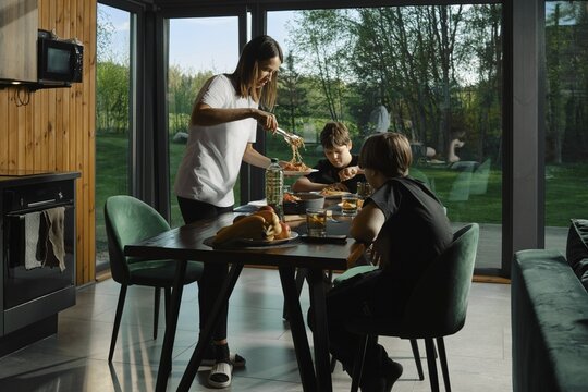 Young mom serves a balanced meal to two children at a dining table in a bright kitchen with large windows. Their warm interaction highlights family bonding while sharing nutritious food