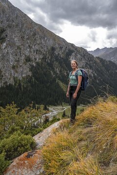 A female hiker stands on a grassy slope against an impressive mountain backdrop under a cloudy sky, Chong Kyzyl Suu Valley, Terskey Ala Too, Tien-Shan Mountains, Kyrgyzstan