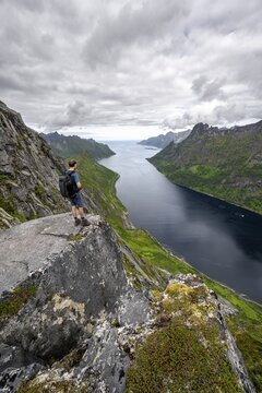 View of &Oslash;yfjorden fjord and mountains, view from Barden mountain, young man standing on a cliff, Senja, Norway