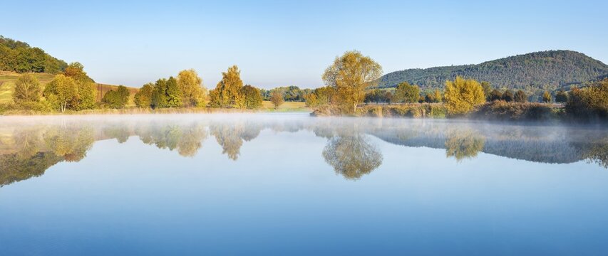 Lake with morning mist in autumn, trees on the shore and hills are reflected, Thuringia, Germany