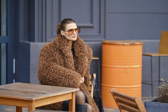 A fashionable woman sits outside a cafe, wearing a large brown fur coat and stylish sunglasses. She appears relaxed as she enjoys the atmosphere in an urban environment during daylight