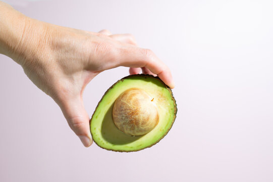 Female hand presenting half an avocado on a light pink background.