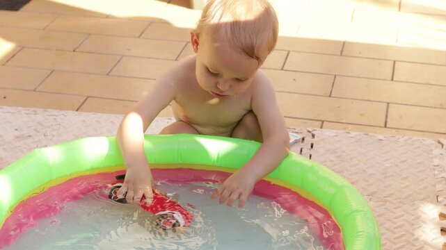 Close up of a cute toddler boy playing with a toy car inside a small inflatable swimming pool on a patio. Little kid splashing water with a plastic truck on a sunny summer day