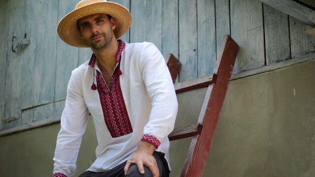 Portrait of smiling guy in straw hat against the background of his house. Young ukrainian man wearing an embroidered shirt looking into camera outdoor. Concept of national identity and patriotism