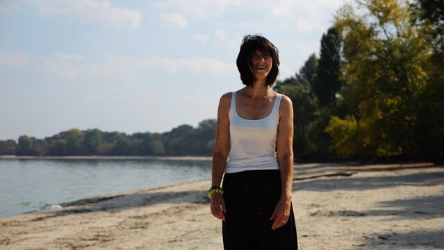 Happy adult woman going on sandy beach near shoreline at sunny day. Beautiful lady walking on seashore with calm waves at background. Concept of summer vacation or holiday. Slow motion