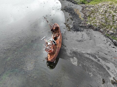 Aerial view of the rusted Stykkisholmur Shipwreck resting on a dark volcanic sand beach near the calm coastal waters in Breidhabolsstadhur, Vesturland, Iceland.