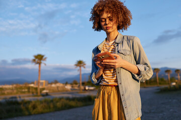 Fototapeta premium Woman on a sunny outdoor street uses smartphone, casual denim shirt over striped tee, flowing skirt, palm trees in background, candid lifestyle moment, modern relaxation