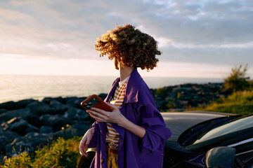 Fototapeta premium A woman stands by a car on the coast at sunset, her hair blowing in the breeze, looking away while holding a tablet. A lifestyle moment of independence and travel.