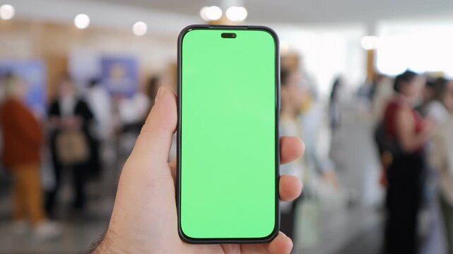 Hand holds phone with green screen. Background shows blurred crowd in indoor space. Suitable for app demos, digital presentations, or UI mockups. Neutral emotion due to blurred faces