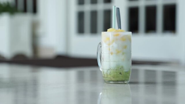 Refreshing tropical fruit dessert drink in a glass with coconut milk, pineapple chunks, and green jelly, served on a table with soft natural light.