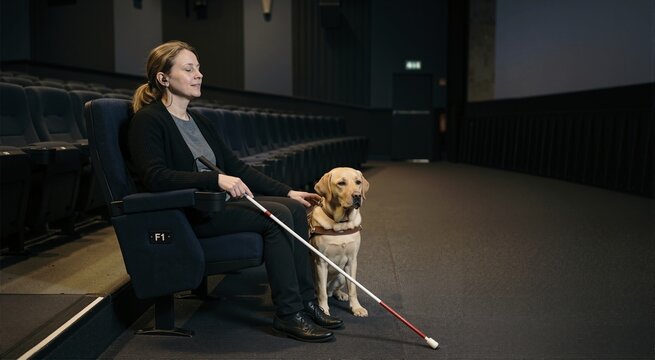 Blind woman with guide dog in movie theater. Visually impaired female listening to audio description with earphones. Accessibility and inclusion concept