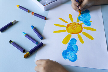 Child's hands drawing a yellow sun and blue clouds with colorful crayons on white paper, scattered markers on light blue table, top view, creativity concept. © Irina