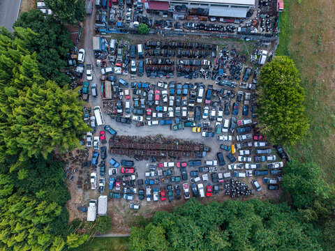 Aerial view of a car scrapyard filled with numerous salvaged vehicles and spare parts, surrounded by dense green trees in Settimo Torinese, Piedmont, Italy.