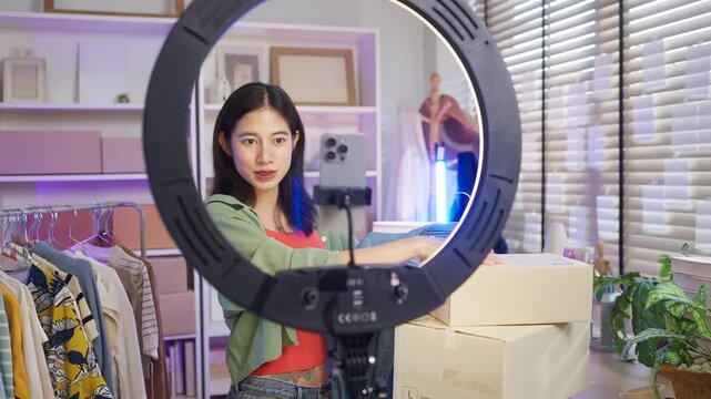 Asian woman live streaming fashion clothing sales with smartphone and ring light, modern retail e-commerce entrepreneur, social commerce influencer and generation z digital side hustle concept