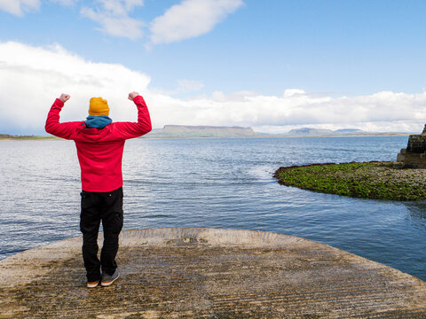 Person in red jacket and yellow hat stands on stone pier. Sea stretches wide under dramatic cloudy sky. Dark storm clouds loom over water horizon. County Sligo, Ireland. Benbulbin mountain viewpoint.