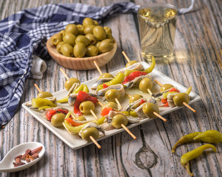 Traditional Spanish gildas skewers with green olives anchovies and hot peppers served on a plate with white wine on a rustic wooden table