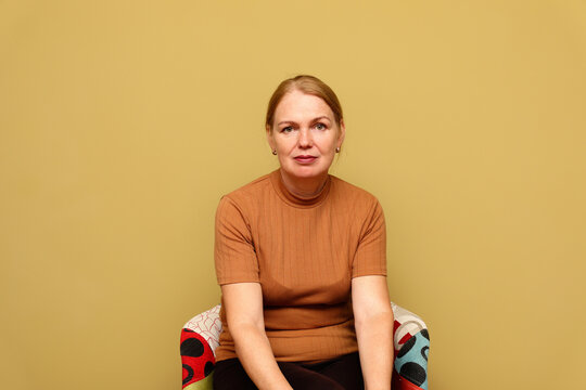 Portrait of mature middle aged woman looking at camera against olive background. elegant lady sitting in colorful armchair, retirement aging concept or mental health template with copy space