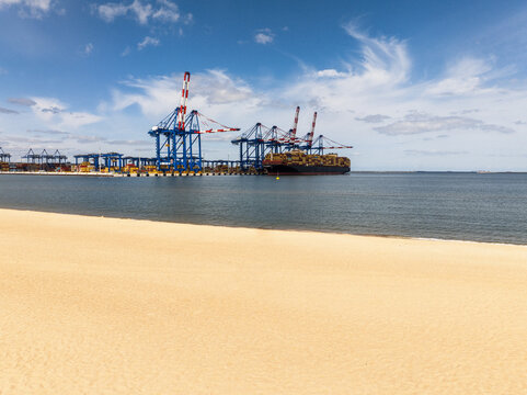 Aerial view of a container terminal with large cranes and a cargo ship docked at the port, seen from a sandy beach in Gdansk, Pomeranian Voivodeship, Poland.