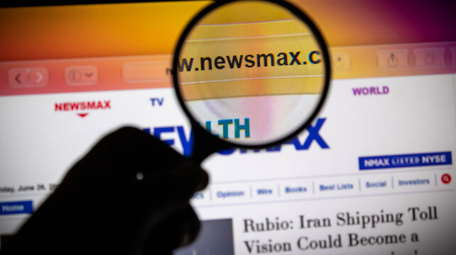 Closeup of a hand holding a magnifying glass over the newsmax website on a computer screen