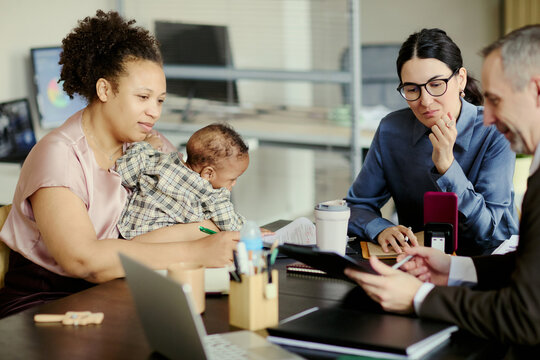 African American young mother holding baby while joining office meeting with colleagues, balancing childcare and career. Ideal for working parent, maternity policy, inclusive workplace marketing