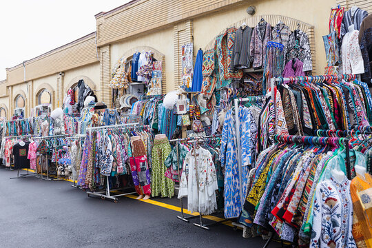 Colorful traditional clothing and textiles displayed on racks outside Siab Bazaar in Samarkand. Vibrant ikat robes, embroidered garments, and scarves create an abundant market scene.