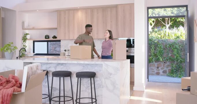 Diverse couple unpacking boxes at marble island, woman entering carrying box, preparing kitchen