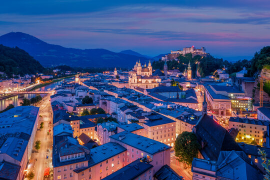 A wide aerial perspective of Salzburg, Austria, featuring the Salzach river flowing past the illuminated historic center and Hohensalzburg Fortress at twilight.