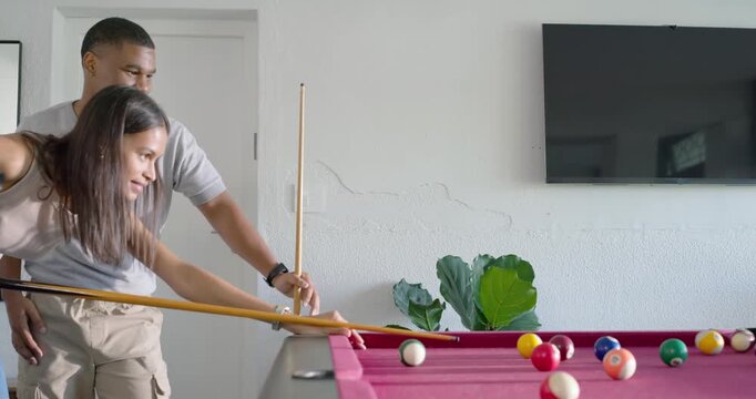 African American couple in home game room, man entering guiding her cue teaching aim, balls rolling