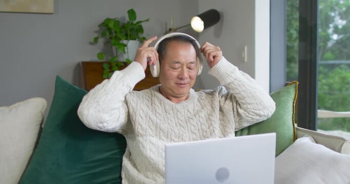 Senior Asian man in sweater reaching up picking white headphones and using laptop at home listening