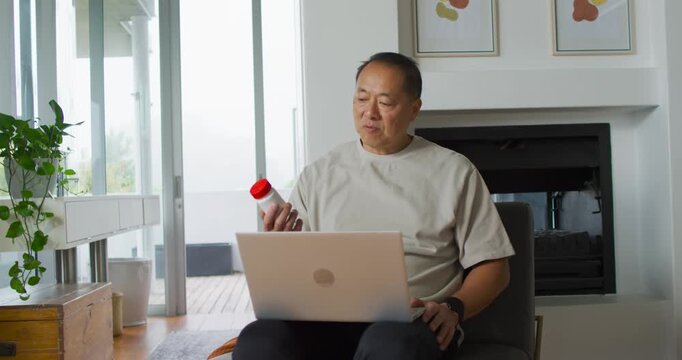 Senior Asian man sitting in living room chair checking bottle after reading laptop, verifying label