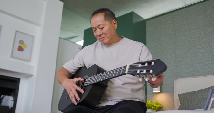 Senior Asian man sitting on bed, plucking strings and adjusting pegs to tune black classical guitar