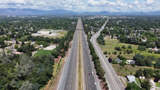 Multi-lane highway and parallel railway split suburban landscape, traffic flowing beneath distant mountain range under partly cloudy blue sky. Redding, California, USA. Aerial drone back wide shot