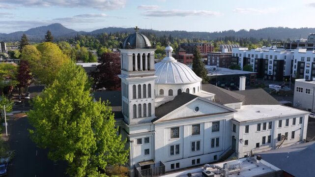 Domed historic courthouse anchors downtown district, bell tower rising above tree-lined streets as warm evening light bathes distant green hills. Eugene, Washington, USA. Aerial drone view.