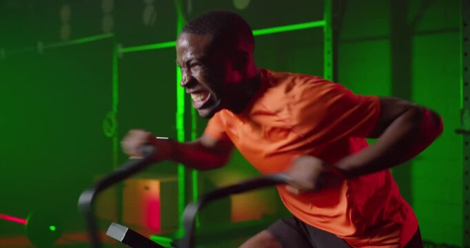 African American man starting assault bike sprint at gym, pedaling hard in orange shirt for fitness