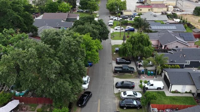 Residential street lined with mature shade trees and parked vehicles, suburban homes featuring solar panels and fenced backyards, sunny day. Stockton, California, USA. Aerial drone flyover shot