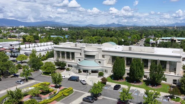Elegant columned civic building surrounded by mature trees and busy parking lots, distant mountains rising beneath bright sunny blue summer sky. Redding, California, USA. Aerial drone approaching shot