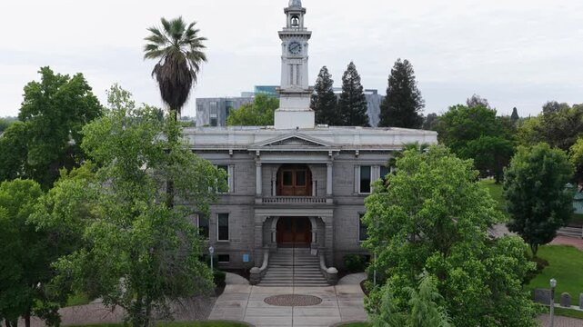 Historic courthouse with clock tower stands behind manicured lawn, grand staircase and carved arched doorways framed by mature trees, cloudy sky. Madera, California, USA. Aerial drone rising shot