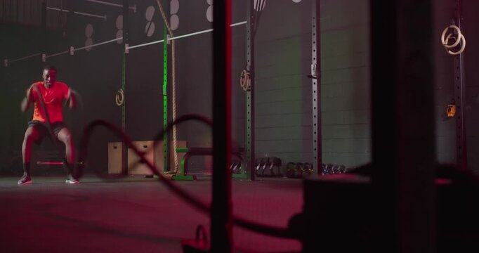 African American man entering left frame, doing squat rope waves in gym for cardio, copy space