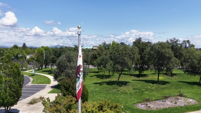 California state flag flutters above grassy park, winding pathway curving through scattered oak trees beneath clear blue sunny summer sky. Redding, California, USA. Aerial drone view.