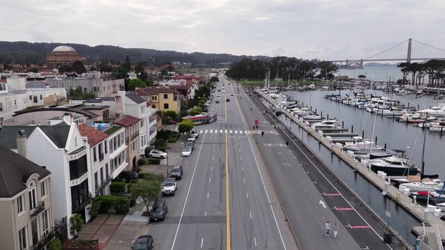 Tree-lined boulevard runs parallel to marina filled with sailboats, Golden Gate Bridge and domed landmark rising along distant horizon. San Francisco, California, USA. Aerial drone flyover shot