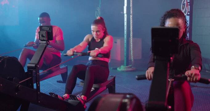 Diverse group rowing on rowers at gym, front woman training, checking trackers, copy space
