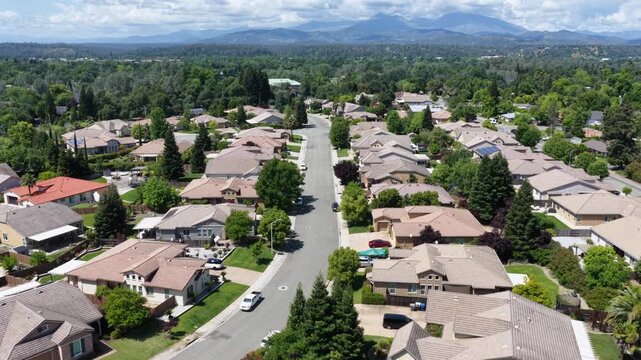 Curved residential street winds past tile-roofed homes and mature trees, distant mountain range rising beneath bright sunny blue summer sky. Redding, California, USA. Aerial drone backwards shot
