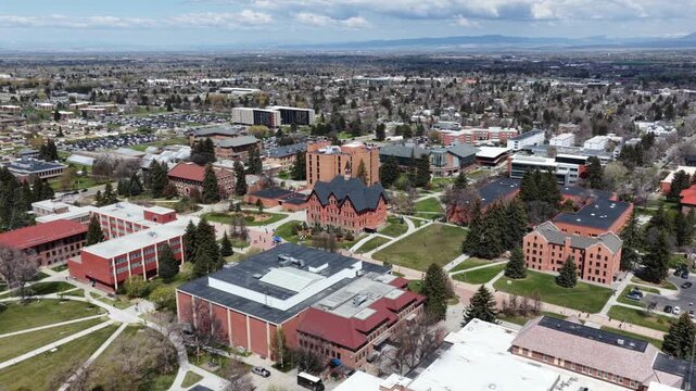 Historic brick university buildings surround tree-lined walkways, students crossing campus quad beneath distant mountains and blue cloudy sky. Bozeman, Montana, USA. Aerial drone rising top down