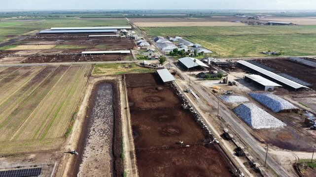 Sprawling Chowchilla farmland reveals cattle feedlot pens, irrigation channels and crop rows stretching toward distant grain silos beneath wide open sky. California, USA. Aerial drone flyover shot