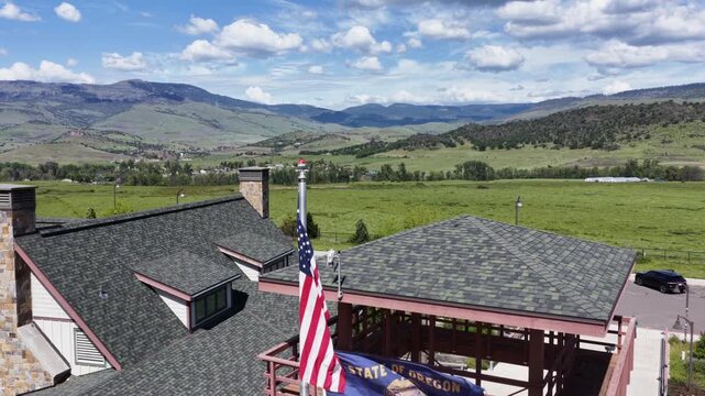 American, Oregon state and POW MIA flags fly above lodge-style welcome center with stone chimney and covered deck, framed by green valley and mountain range. Oregon, USA. Aerial drone view.