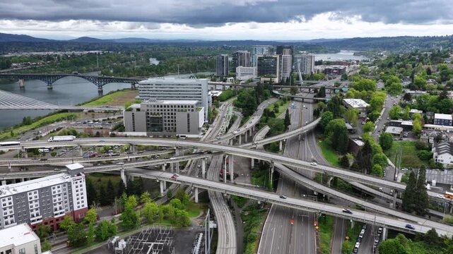 Highway interchange weaves beneath stormy clouds near riverfront, traffic and school bus crossing ramps flanked by modern towers and green hillside. Portland, Oregon, USA. Aerial drone flyover shot