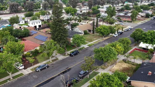 Tree-lined residential street showcases single-family homes with solar panels, parked cars along curb beneath mature shade trees and rooftops. Madera, California, USA. Aerial drone flyover shot