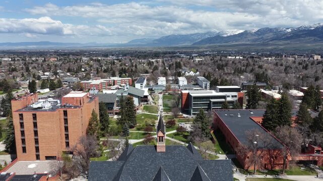 Steep gabled roofline and spire crown historic university hall, students crossing pathways near brick dormitory tower beneath snow-capped mountains. Bozeman, Montana, USA. Aerial drone rising wide fly